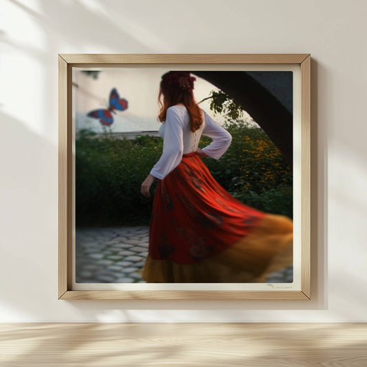 Eine Frau in folkloristischer Kleidung tanzt auf Kopfsteinpflaster durch einen Steinbogen, im Hintergrund ein großer Schmetterling am Meer.. Rahmen natur 30x30