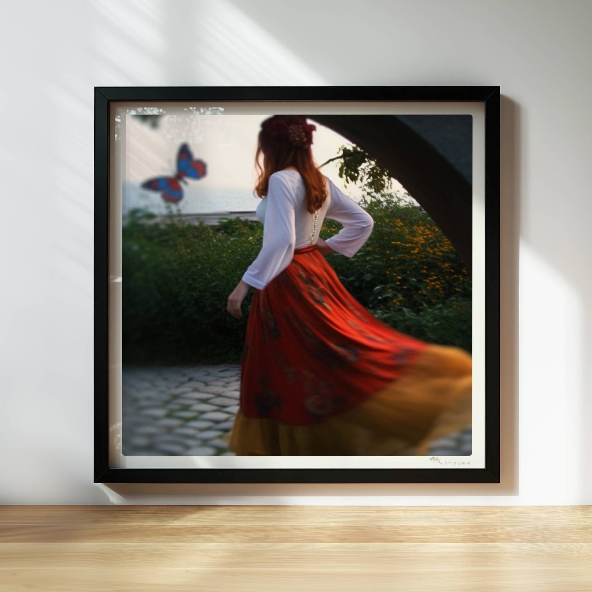 Eine Frau in folkloristischer Kleidung tanzt auf Kopfsteinpflaster durch einen Steinbogen, im Hintergrund ein großer Schmetterling am Meer.. Rahmen schwarz 30x30