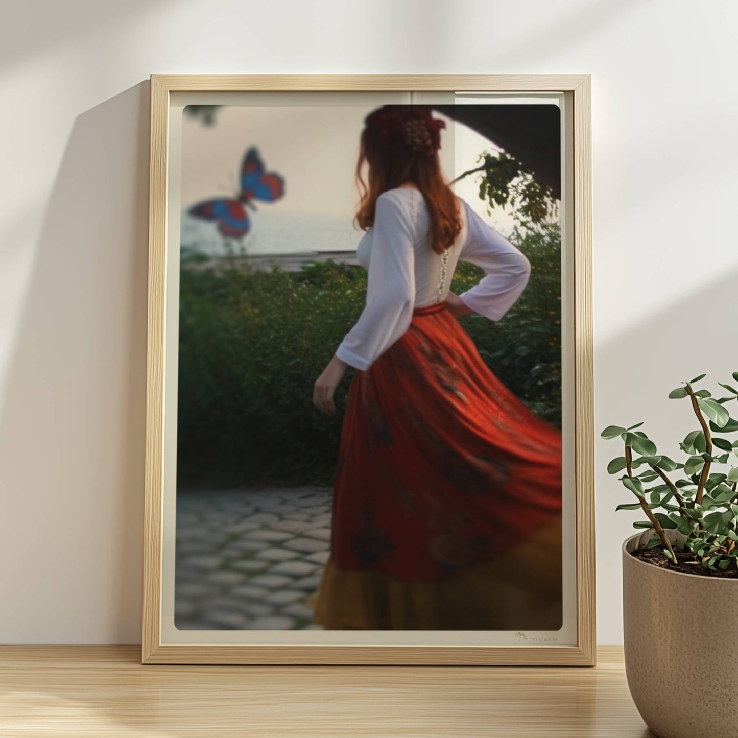 Eine Frau in folkloristischer Kleidung tanzt auf Kopfsteinpflaster durch einen Steinbogen, im Hintergrund ein großer Schmetterling am Meer.. Rahmen natur 30x40