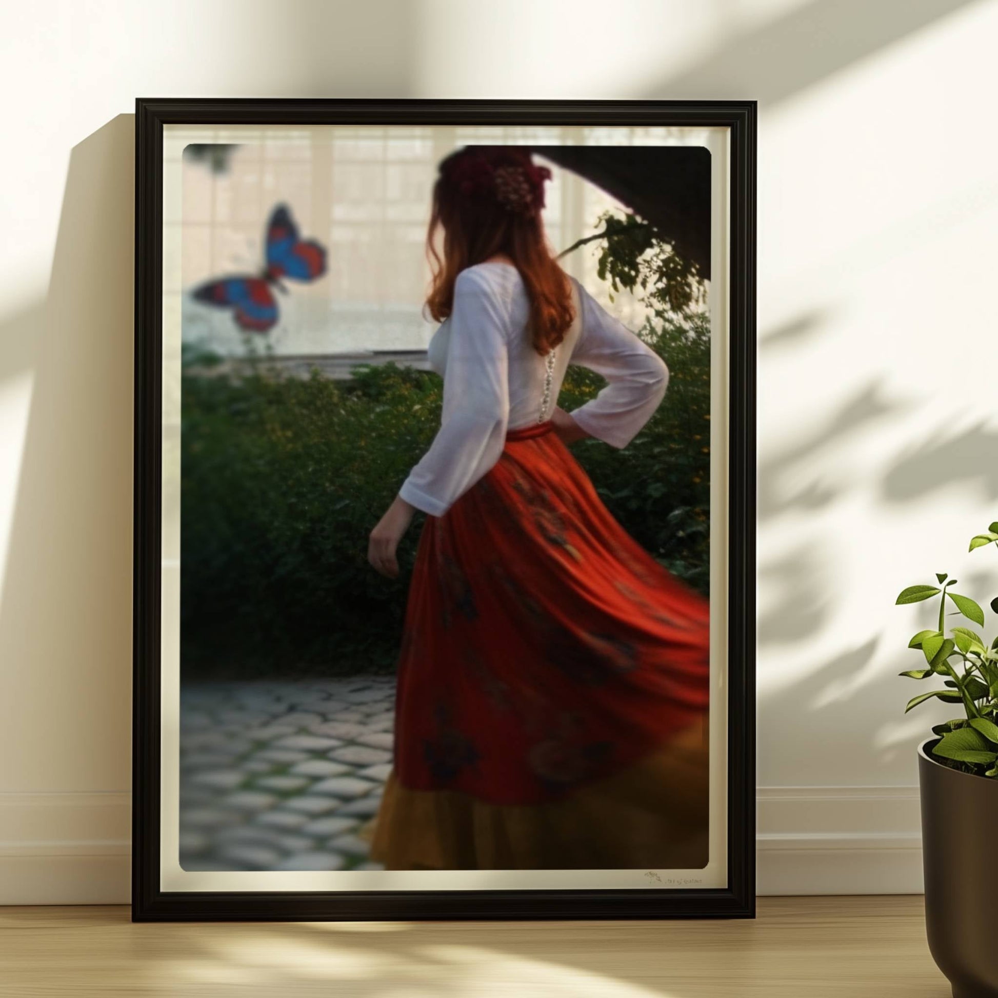 Eine Frau in folkloristischer Kleidung tanzt auf Kopfsteinpflaster durch einen Steinbogen, im Hintergrund ein großer Schmetterling am Meer.. Rahmen schwarz 30x40