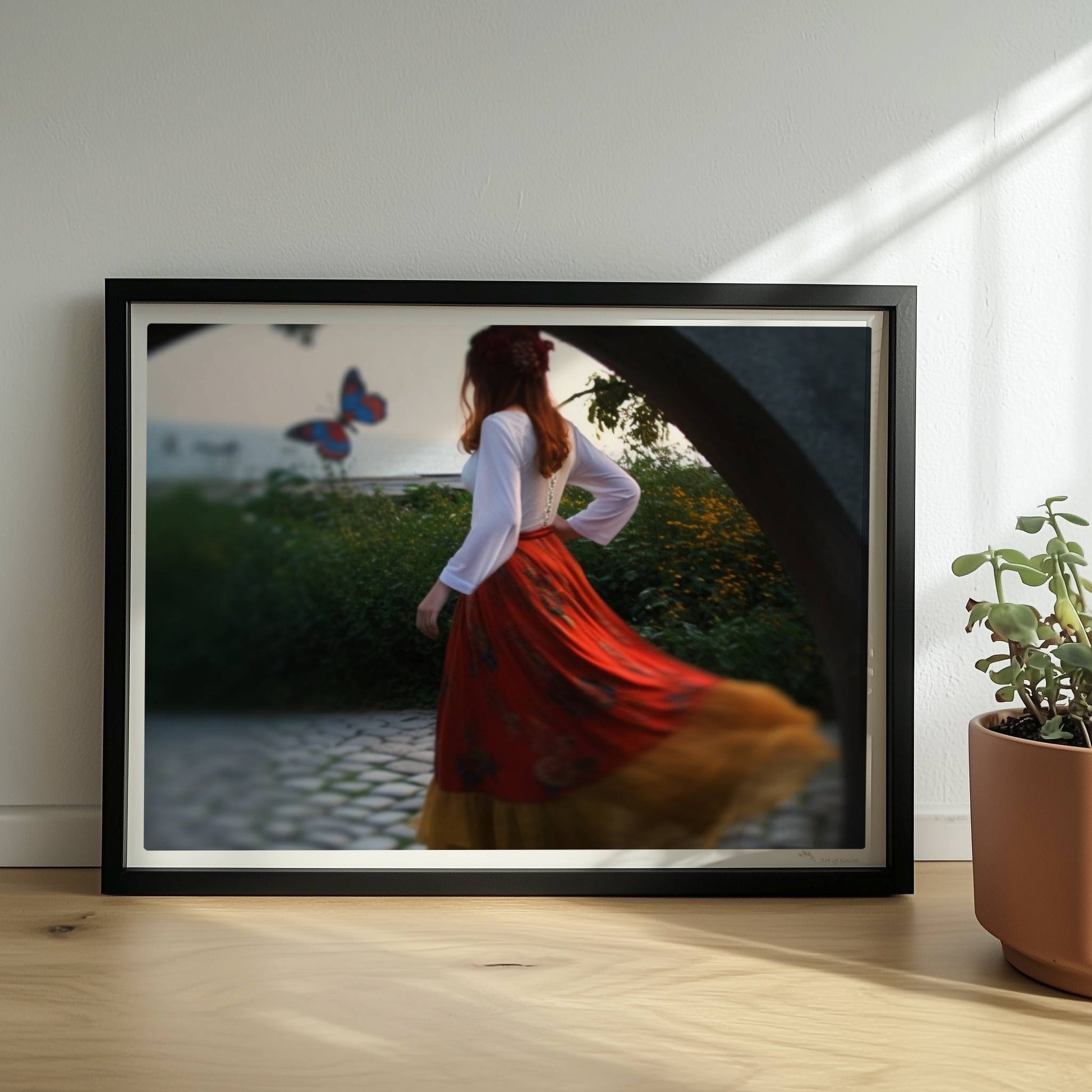 Eine Frau in folkloristischer Kleidung tanzt auf Kopfsteinpflaster durch einen Steinbogen, im Hintergrund ein großer Schmetterling am Meer.. Rahmen schwarz 40x30