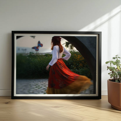Eine Frau in folkloristischer Kleidung tanzt auf Kopfsteinpflaster durch einen Steinbogen, im Hintergrund ein großer Schmetterling am Meer.. Rahmen schwarz 40x30