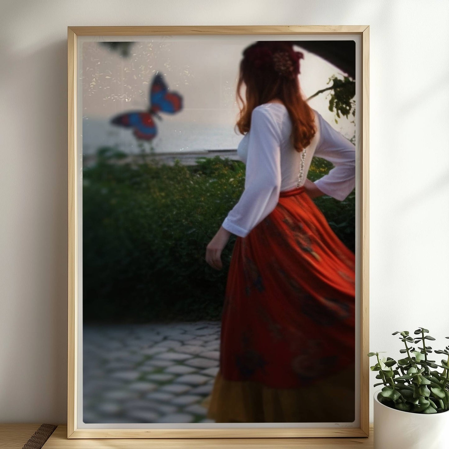 Eine Frau in folkloristischer Kleidung tanzt auf Kopfsteinpflaster durch einen Steinbogen, im Hintergrund ein großer Schmetterling am Meer.. Rahmen natur 50x70