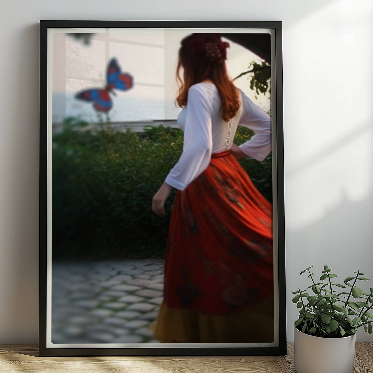 Eine Frau in folkloristischer Kleidung tanzt auf Kopfsteinpflaster durch einen Steinbogen, im Hintergrund ein großer Schmetterling am Meer.. Rahmen schwarz 50x70