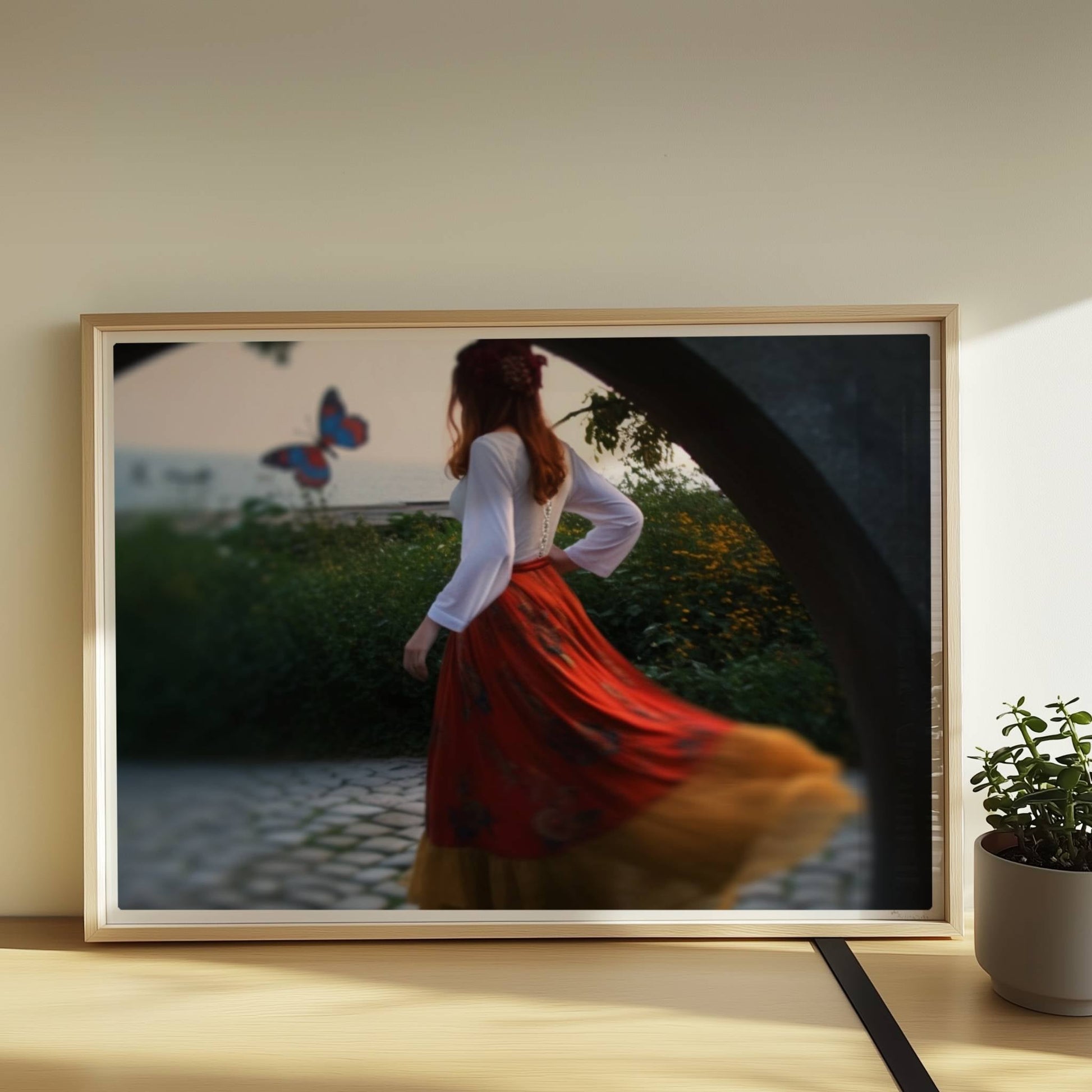 Eine Frau in folkloristischer Kleidung tanzt auf Kopfsteinpflaster durch einen Steinbogen, im Hintergrund ein großer Schmetterling am Meer.. Rahmen natur 70x50