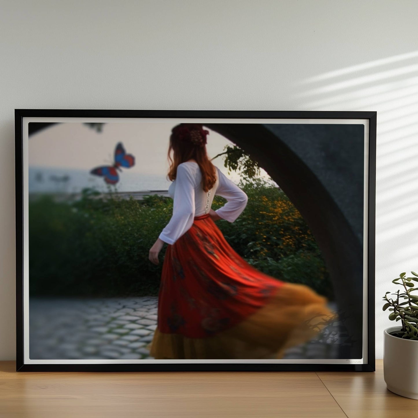 Eine Frau in folkloristischer Kleidung tanzt auf Kopfsteinpflaster durch einen Steinbogen, im Hintergrund ein großer Schmetterling am Meer.. Rahmen schwarz 70x50